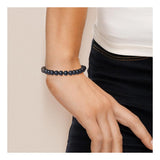 Une personne porte le bracelet Perlinea NOELYSE | Argent & Perles Noires (7/8 mm, fermoir mousqueton) au poignet, la main sur la hanche, vêtue d'un haut blanc et d'un pantalon foncé.