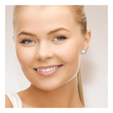 Une femme blonde avec une queue de cheval sourit à l'appareil photo, portant les Boucles d'Oreilles COROLLE | Perles Blanches de Perlinea et un haut blanc. Son maquillage est subtil, ses sourcils sont bien dessinés et l'arrière-plan est sobre.