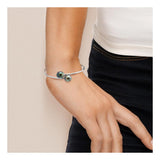Un gros plan montre un poignet portant le bracelet Perlinea Jonc KATALÏA | Perles de Tahiti, un bracelet élégant en argent avec deux perles rondes de Tahiti vert foncé. La personne est vêtue d'un haut blanc et d'un pantalon noir.
