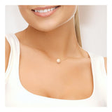 Une femme en haut blanc sourit, mettant en valeur le Perlinea Collier SOFIA | Perle Rose Naturel - un collier délicat avec un seul pendentif en perle naturelle rose reposant sur sa clavicule sur un fond uni et clair.