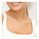 Une femme vêtue d'un haut blanc sourit doucement, portant le Perlinea Collier Cravate avec une Perle de Culture Dorée pendante.